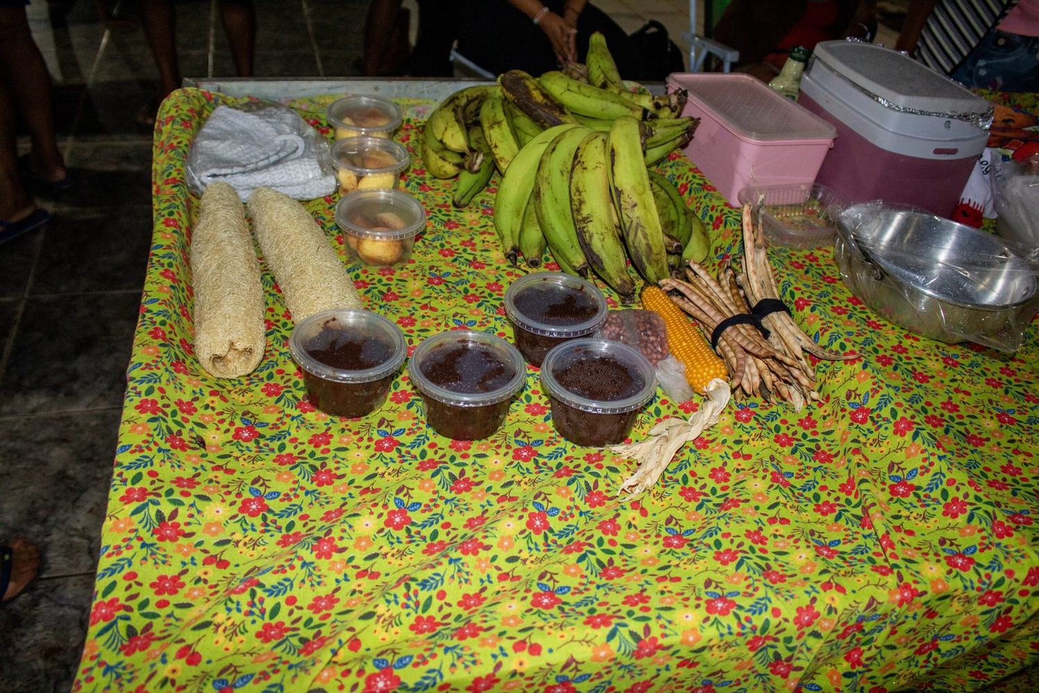Banca com cachos de banana, espigas de milho, potes de doce e bucha vegetal sobre toalha florida