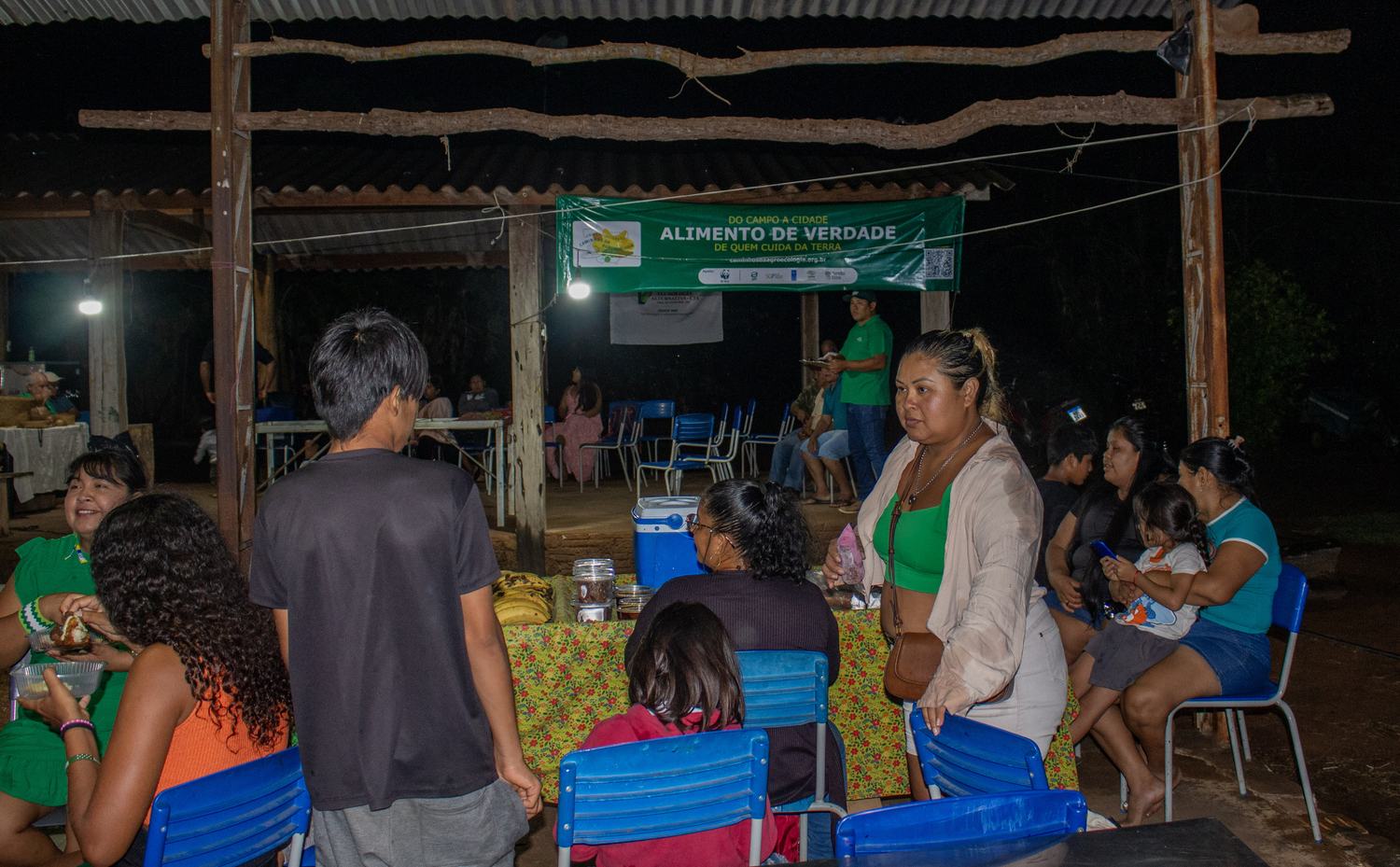 Comunidade reunida no salão da APIC à noite, com as bancas e a faixa da Rota ao fundo