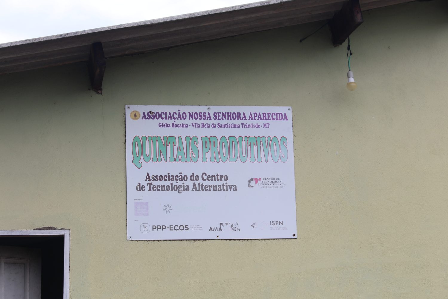 Placa do projeto Quintais Produtivos da Associação Nossa Senhora Aparecida e do CTA, com as logos do PPP-ECOS e do ISPN, na parede do centro social
