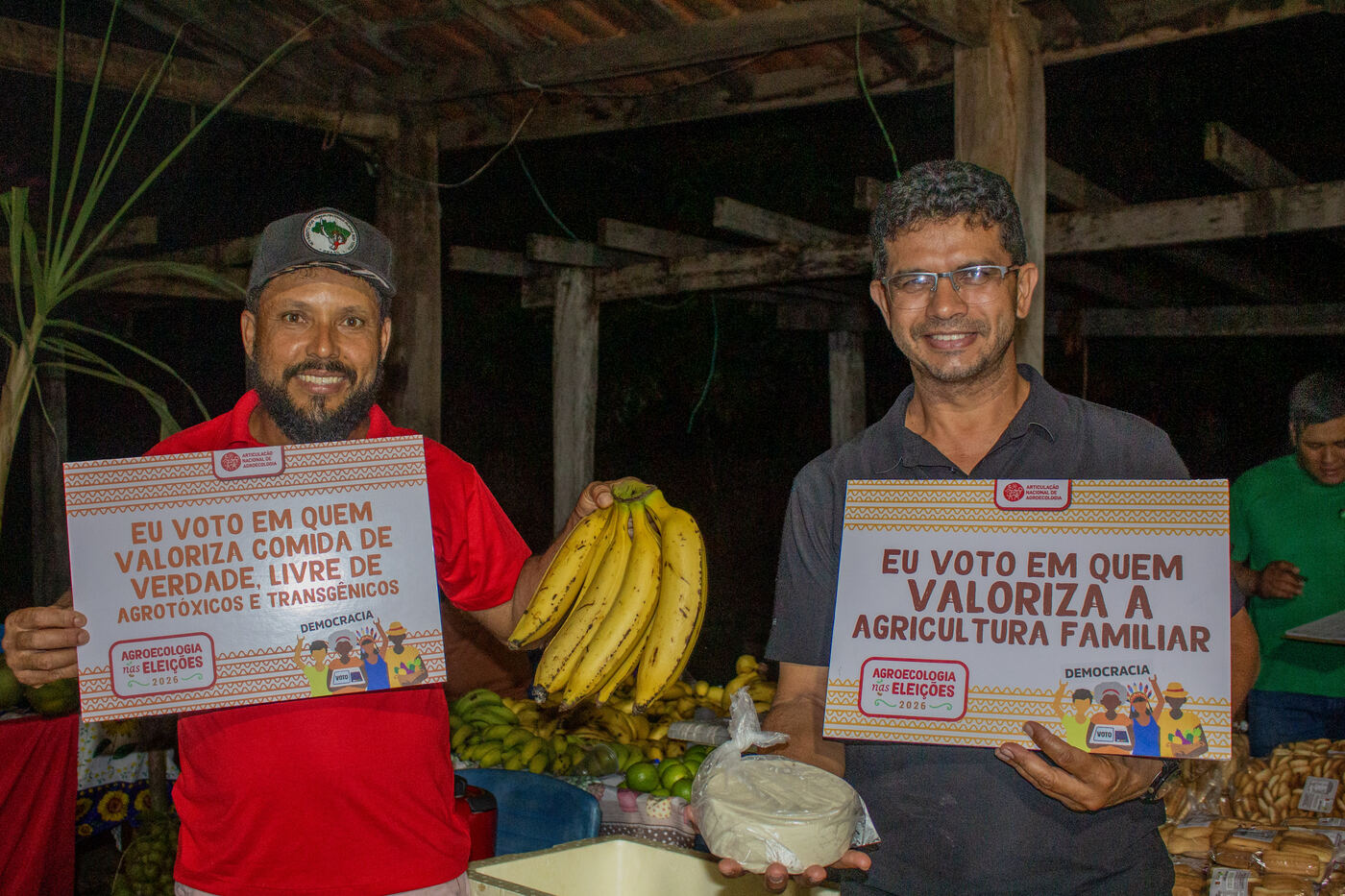 Dois agricultores com placas da campanha Agroecologia nas Eleições na banca de bananas e queijos