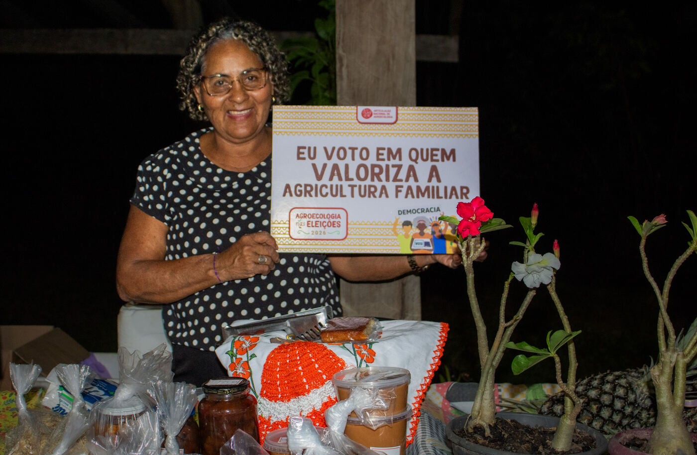 Agricultora na banca de conservas e mudas com placa da campanha