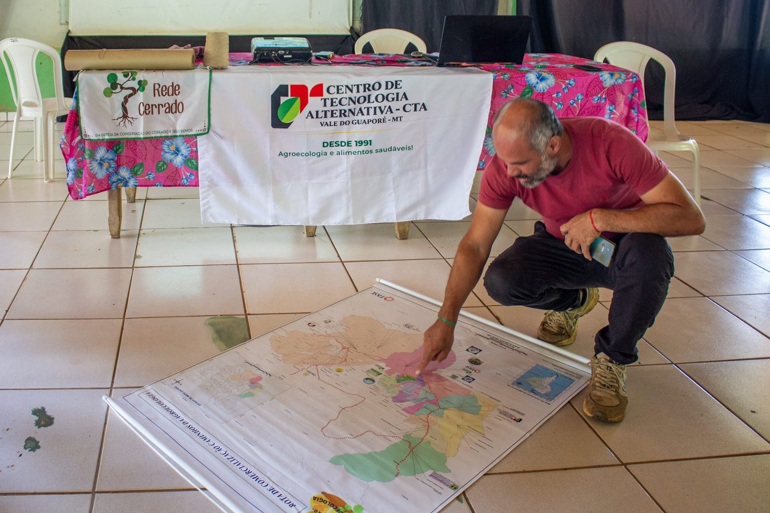 Saguio Moreira, presidente da Coopervales, agachado ao lado do mapa da Rota Caminhos da Agroecologia estendido no chão do salão